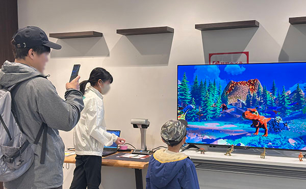 色を塗った3Dの恐竜が映るモニター、それを見ている子ども、写真を撮るお父さん、そばにスタッフがいる様子