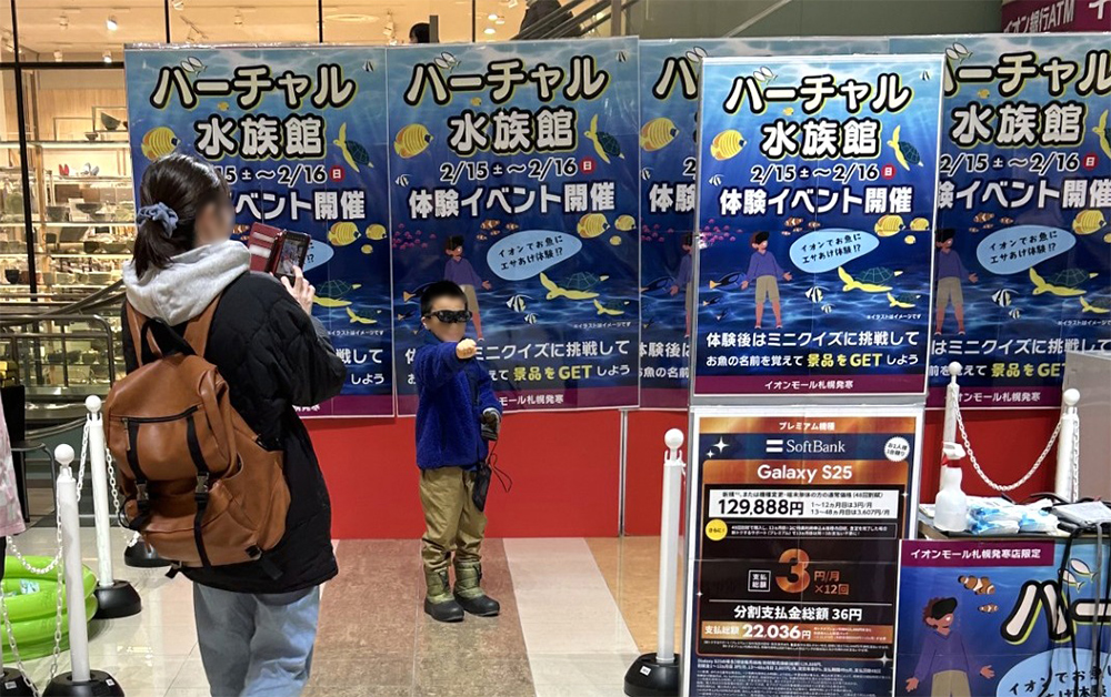 バーチャル水族館と書かれたポスターが並ぶ会場で、黒いスマートグラスをかけて手をグーにして前に出している子どもと写真を撮るお母さん