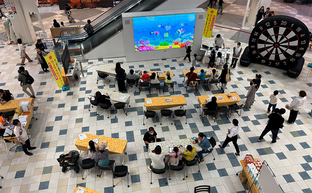 テーブルと椅子が並んだ会場でぬりえをするこどもたちと見守るおとな
