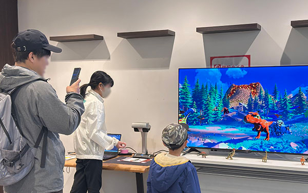 色を塗った3Dの恐竜が映るモニター、それを見ている子ども、写真を撮るお父さん、そばにスタッフがいる様子